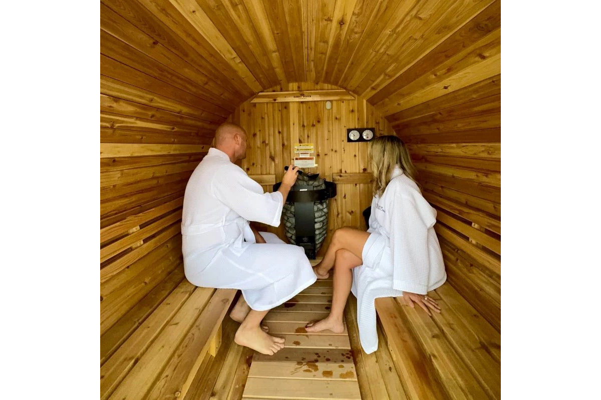 Two people wearing white waffle robes inside wooden barrel sauna with cedar interior, benches, and control panel