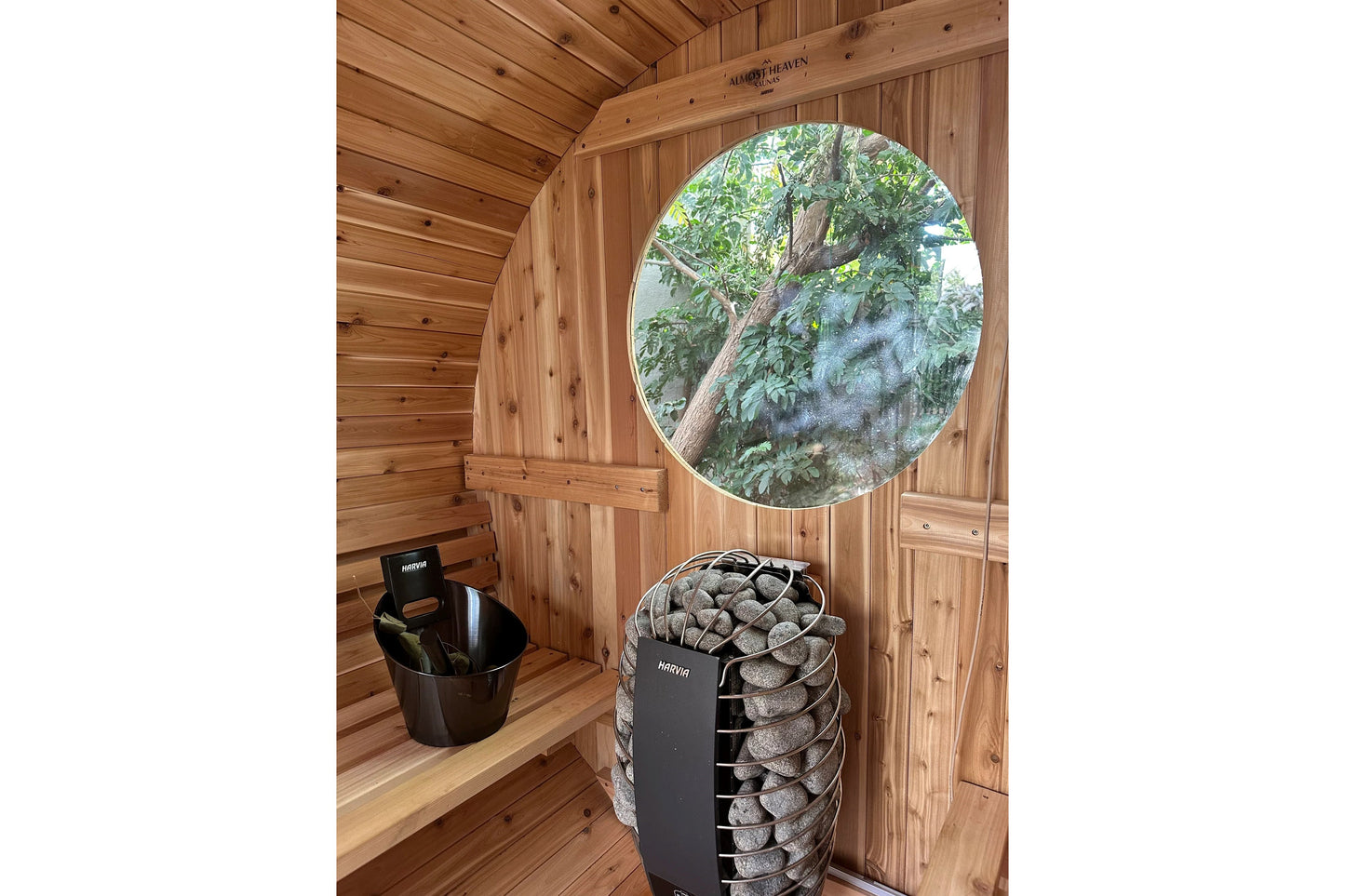 Wood barrel sauna interior with round window, black heater stove filled with stones, and wooden benches