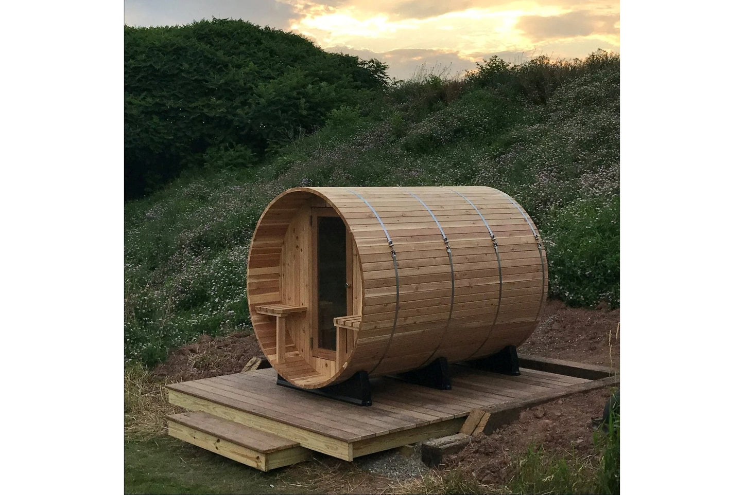 Cylindrical wooden barrel sauna with arched roof, metal bands, entrance door, and wooden deck platform in outdoor garden sett