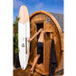 Man holding white and natural wood surfboard next to barrel sauna with open wooden door, outdoor setting