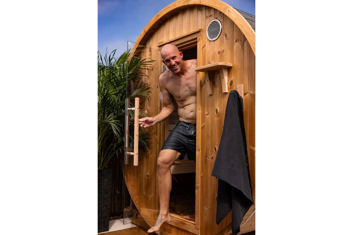 Man exiting wooden barrel sauna with arched roof, round ventilation window, and black towel hanging outside