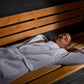 Man lying on wooden bench inside sauna wearing light blue waffle-weave robe