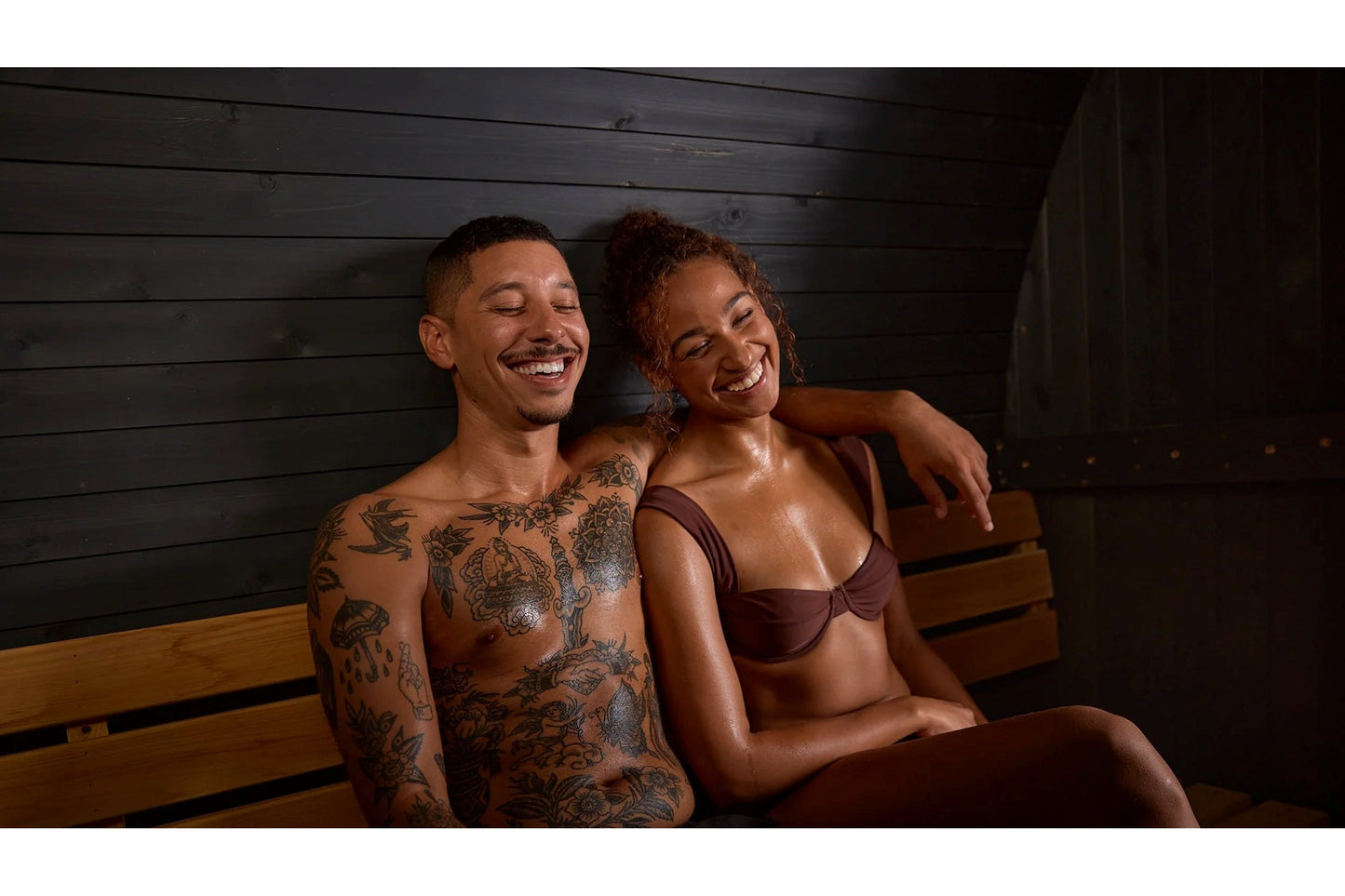 Two people sitting together on wooden bench inside sauna with black shiplap walls