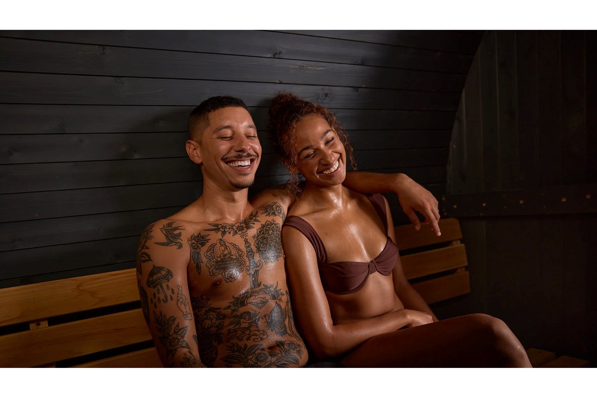 Two people sitting together on wooden bench inside sauna with black shiplap walls