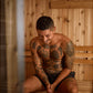 Shirtless man with full-body tattoos sitting in wooden sauna interior with pine walls