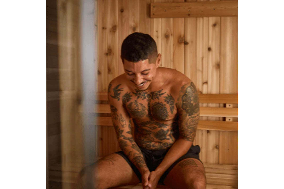 Shirtless man with full-body tattoos sitting in wooden sauna interior with pine walls