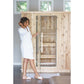 Woman in white robe opening glass door of natural wood sauna with black frame, standing on mat in bathroom setting