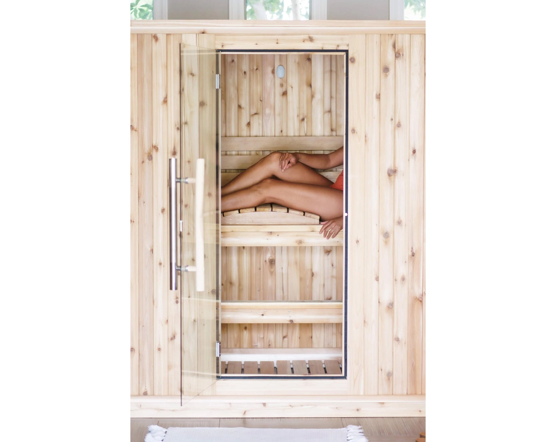 Light wood sauna with glass door and metal frame, person relaxing inside on wooden benches, overhead view