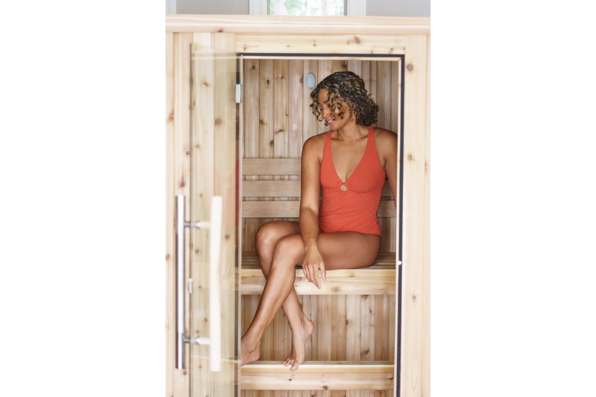 Model wearing coral red one-piece swimsuit sitting inside wooden sauna with glass door and wooden benches