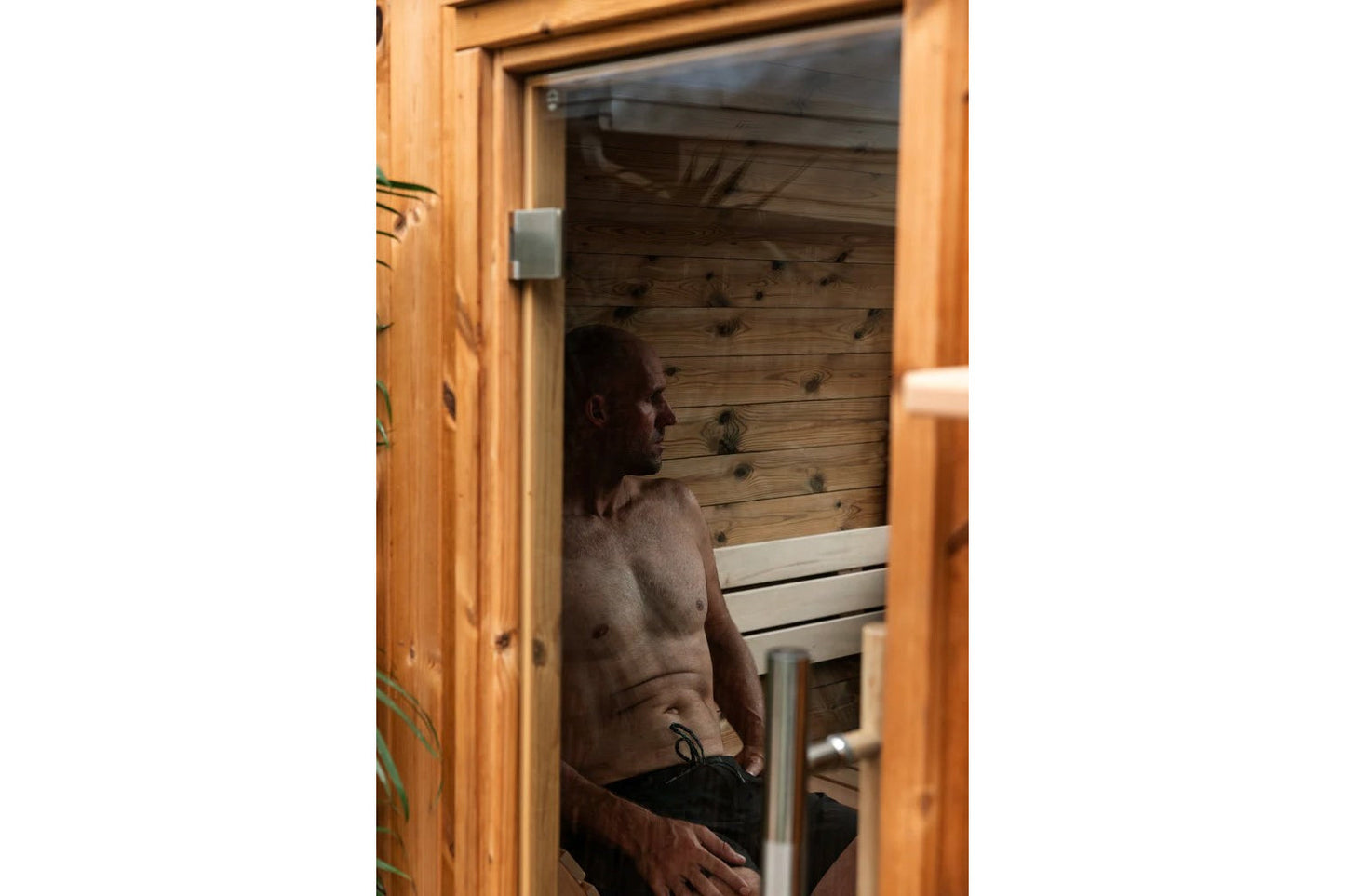 Man sitting inside wooden barrel sauna with glass window and wood interior, viewed through open door