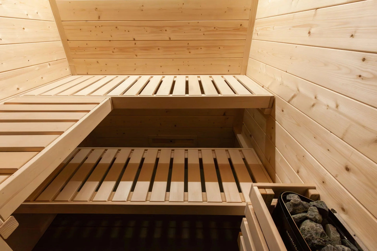 Interior view of wooden sauna with two bench levels, light pine construction, and rock heater basket in corner
