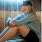 Woman wearing gray long-sleeve top and black shorts sitting inside wooden sauna with blue lighting