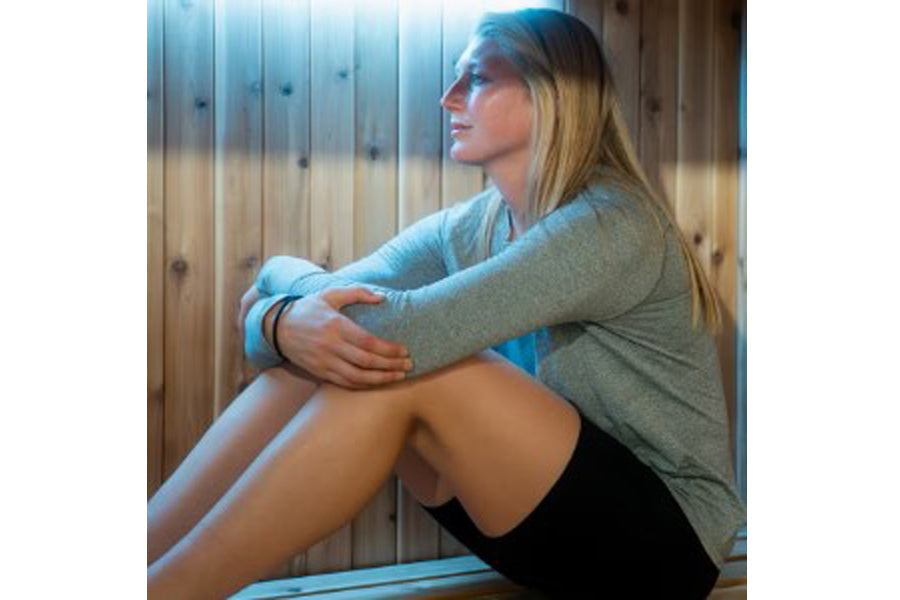 Woman wearing gray long-sleeve top and black shorts sitting inside wooden sauna with blue lighting