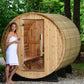 Wooden barrel sauna with curved cedar exterior, open door showing interior benches, model in white towel standing outside, su