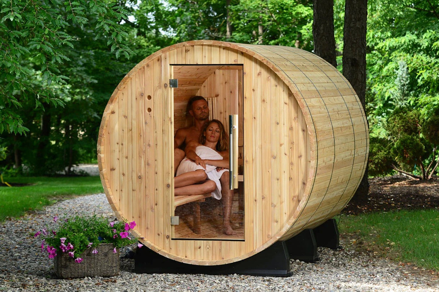 Cylindrical wooden barrel sauna with natural pine construction, open door showing two people inside, set in wooded garden set