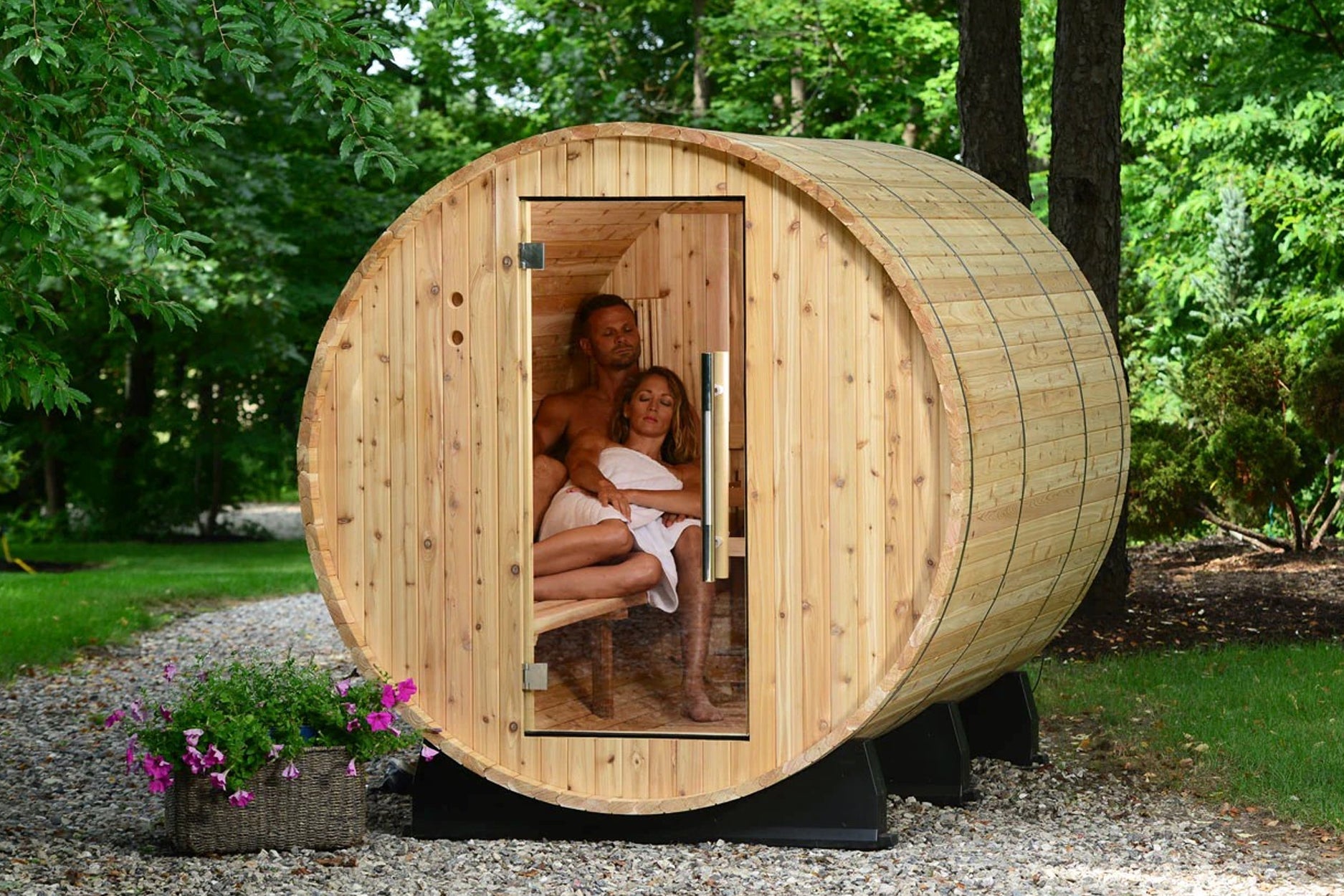 Cylindrical wooden barrel sauna with natural pine construction, open door showing two people inside, set in wooded garden set