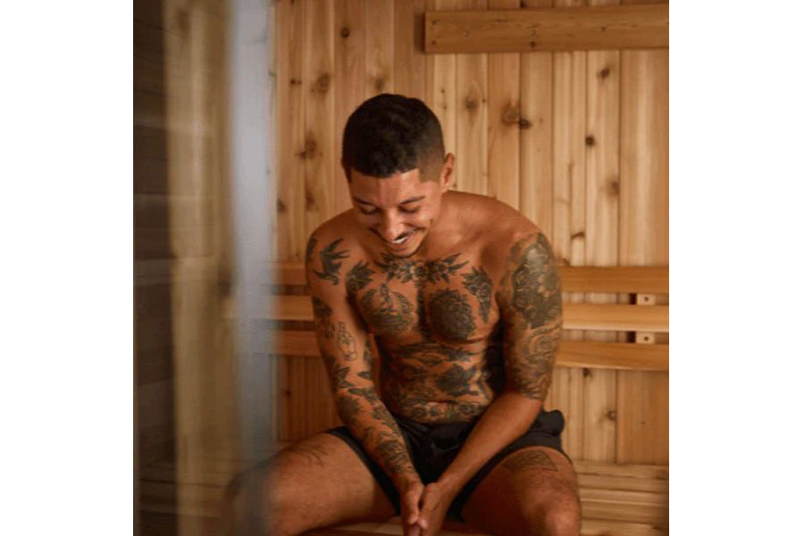 Man sitting in wooden sauna interior with tattoos on chest and arms, wearing black shorts