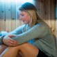 Woman wearing gray long-sleeve top seated inside wooden sauna with blue lighting, demonstrating relaxed posture