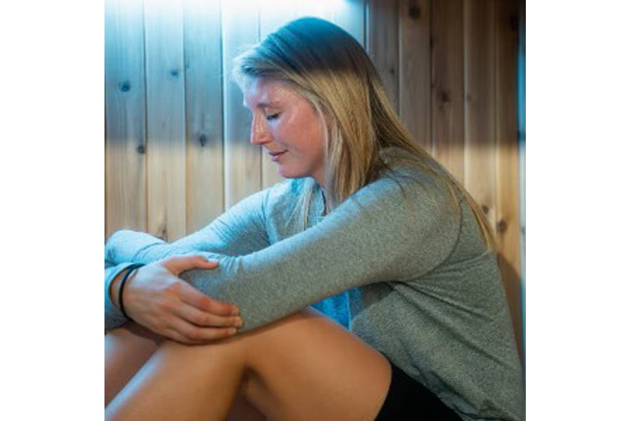 Woman wearing gray long-sleeve top seated inside wooden sauna with blue lighting, demonstrating relaxed posture