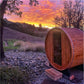 Wooden barrel sauna with open door in rural landscape at sunset, surrounded by gravel and grass with rolling hills in backgro