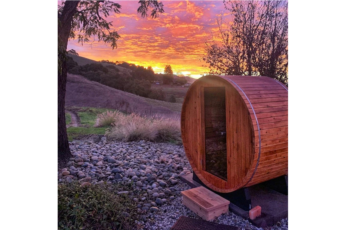 Wooden barrel sauna with open door in rural landscape at sunset, surrounded by gravel and grass with rolling hills in backgro