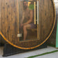 Wooden barrel sauna with glass door, man inside wearing green shorts, gold metal bands around exterior, on concrete pad