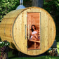 Wooden barrel sauna with open door showing woman seated inside on bench, surrounded by green trees and landscaping