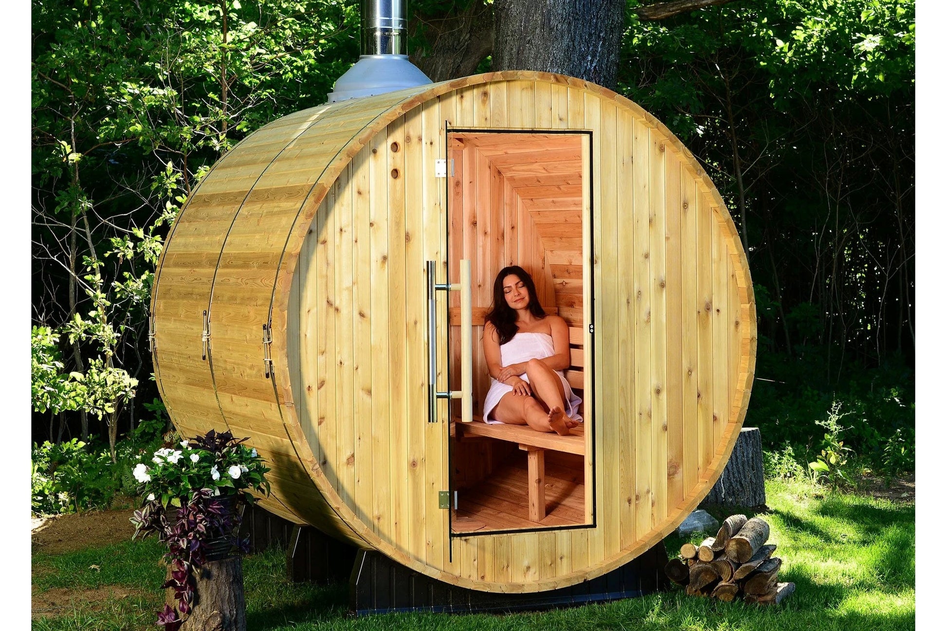 Wooden barrel sauna with open door showing woman seated inside on bench, surrounded by green trees and landscaping