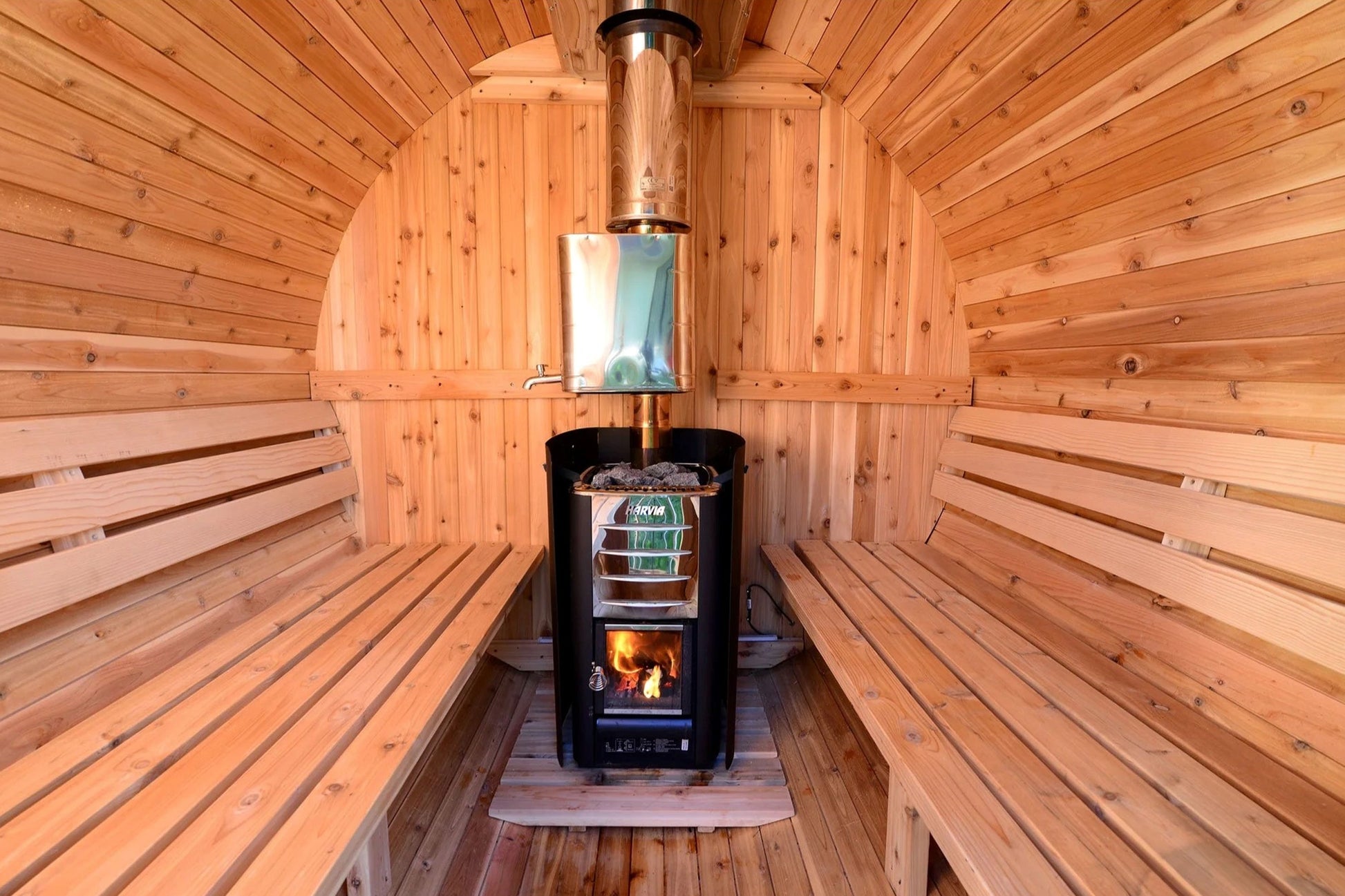 Almost Heaven Seneca barrel sauna interior with wood benches, black wood-burning stove with active fire, and metal chimney pi
