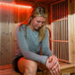 Woman wearing gray long-sleeve shirt inside wooden sauna with red neon lighting, seated on bench