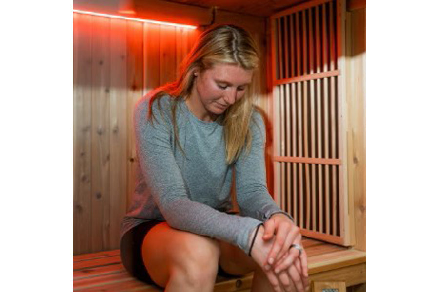 Woman wearing gray long-sleeve shirt inside wooden sauna with red neon lighting, seated on bench