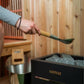 Hand holding wooden-handled sauna scoop with green metal bowl over Harvia sauna bucket filled with stones in wood-lined sauna