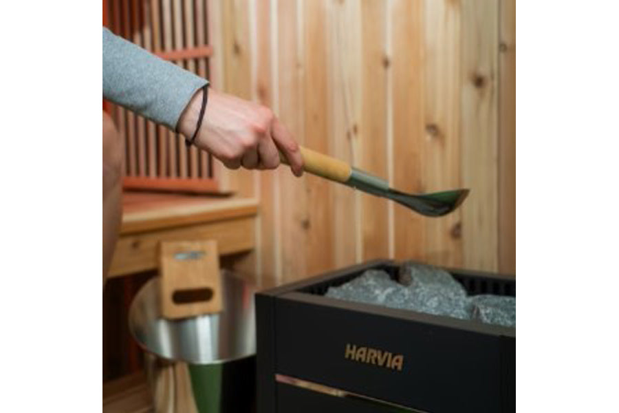 Hand holding wooden-handled sauna scoop with green metal bowl over Harvia sauna bucket filled with stones in wood-lined sauna