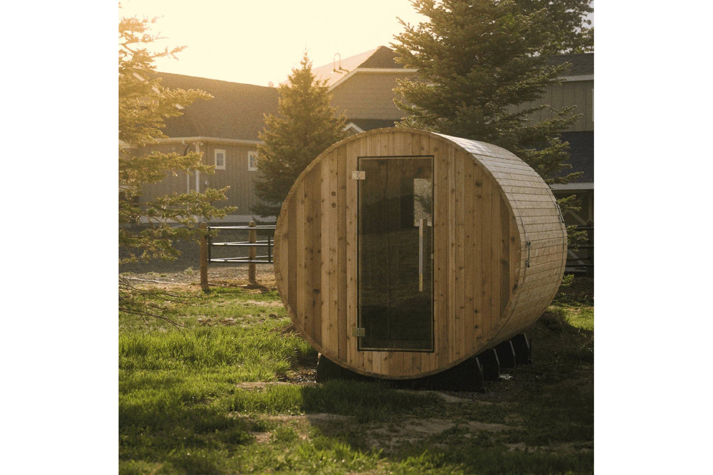 Wooden barrel sauna with rounded cylindrical design and dark glass door, positioned in backyard setting with residential buil