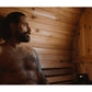 Bearded man with visible tattoos relaxing shirtless inside wooden barrel sauna with horizontal wood paneling