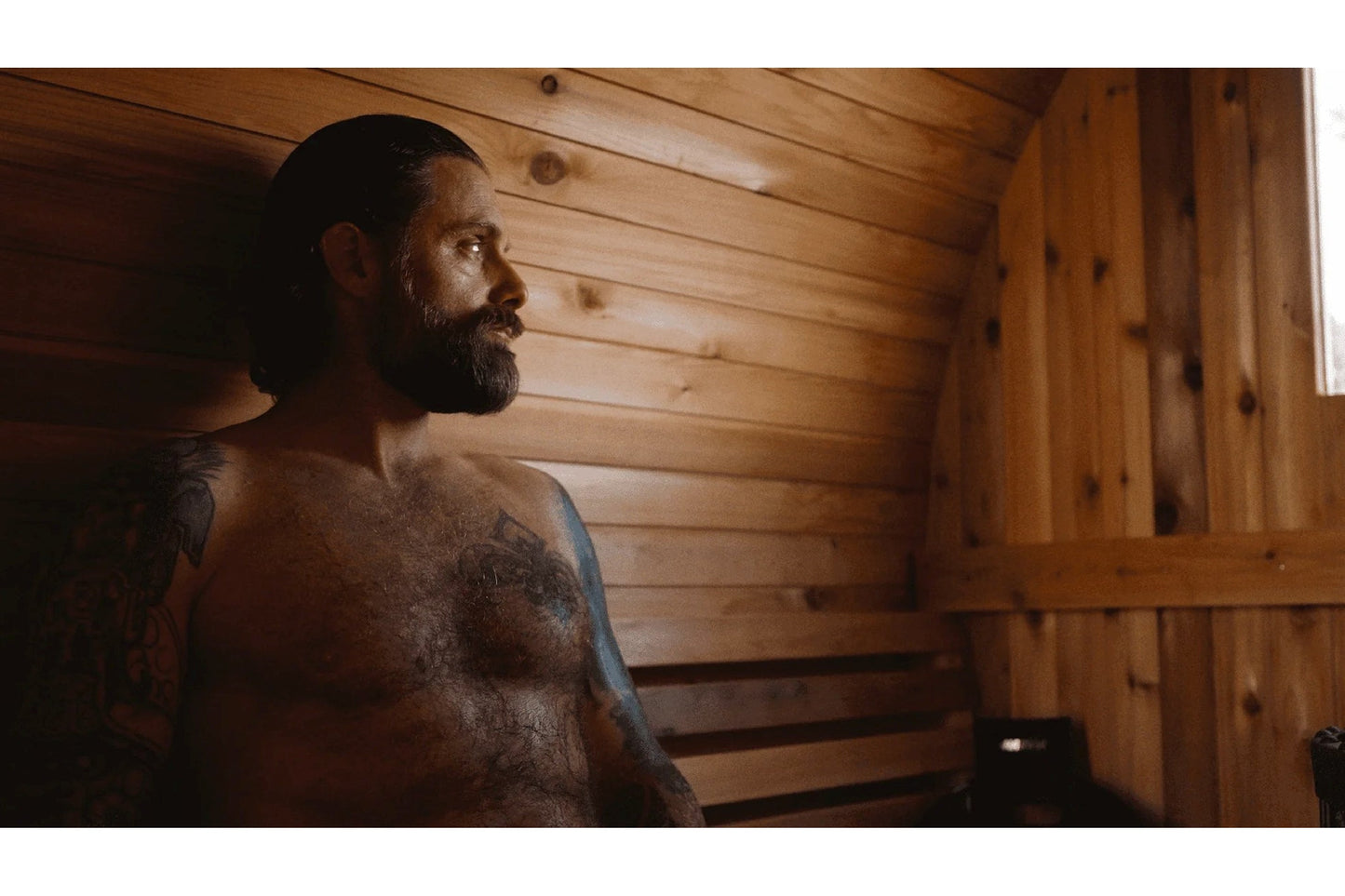 Bearded man with visible tattoos relaxing shirtless inside wooden barrel sauna with horizontal wood paneling
