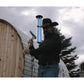 Man in black cowboy hat and jacket working on blue chimney pipe of wooden barrel sauna with metal roofing