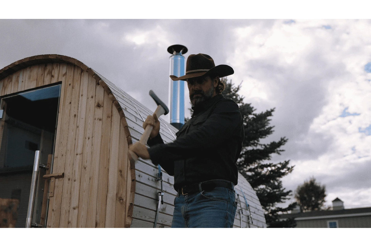 Man in black cowboy hat and jacket working on blue chimney pipe of wooden barrel sauna with metal roofing