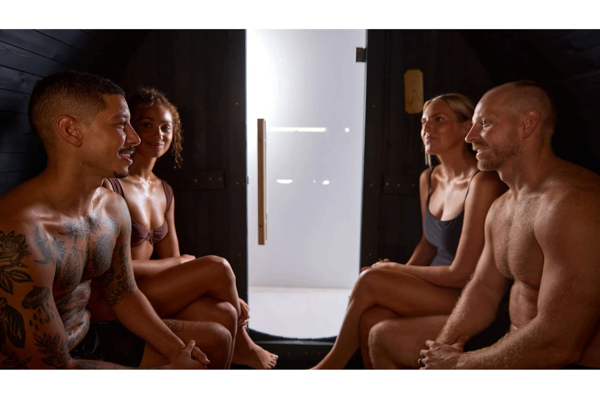 Four people relaxing inside a wooden barrel sauna with glass door and black interior walls