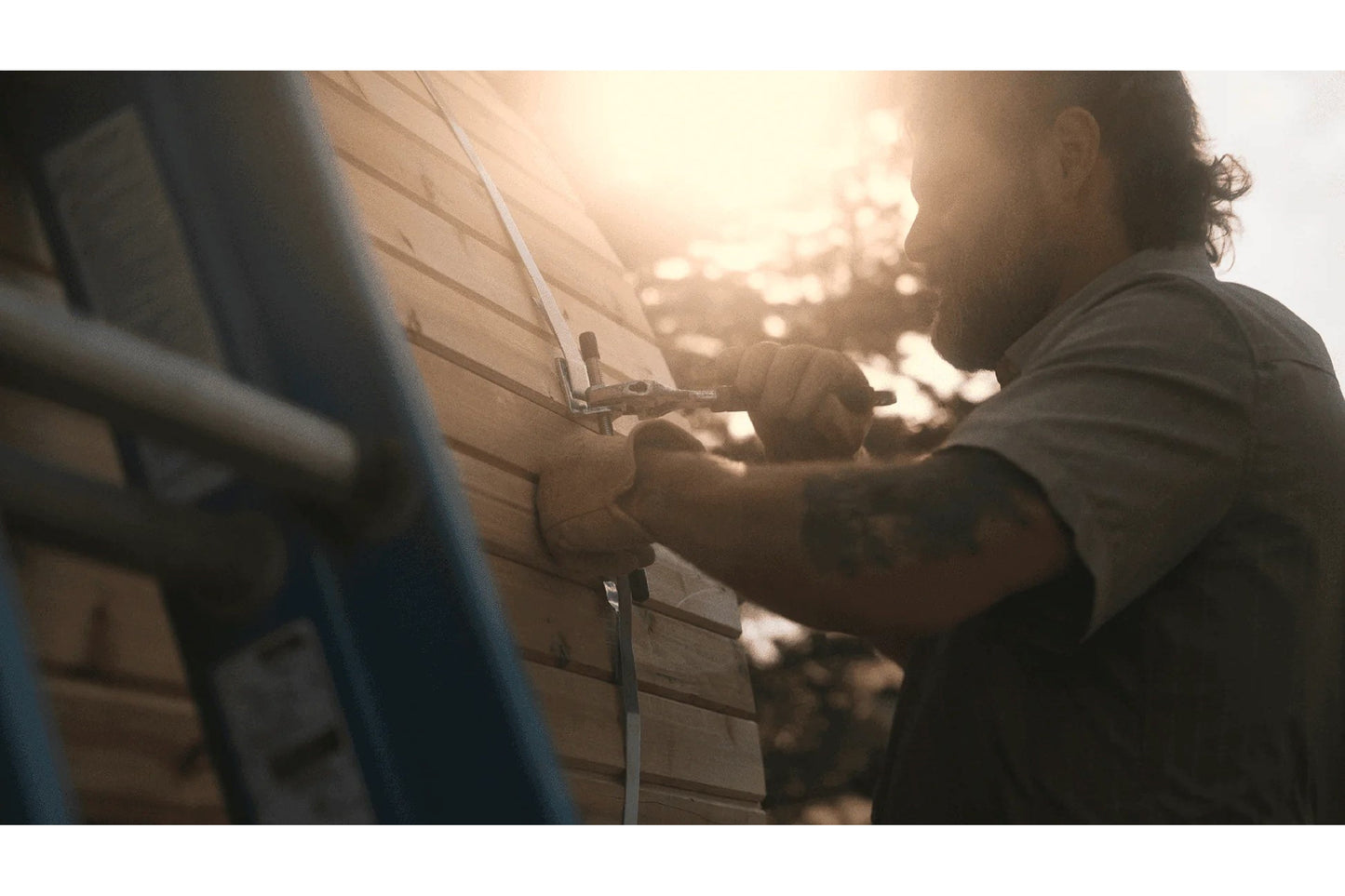 Man in gray shirt installing stainless steel bands on wooden barrel sauna in sunlight