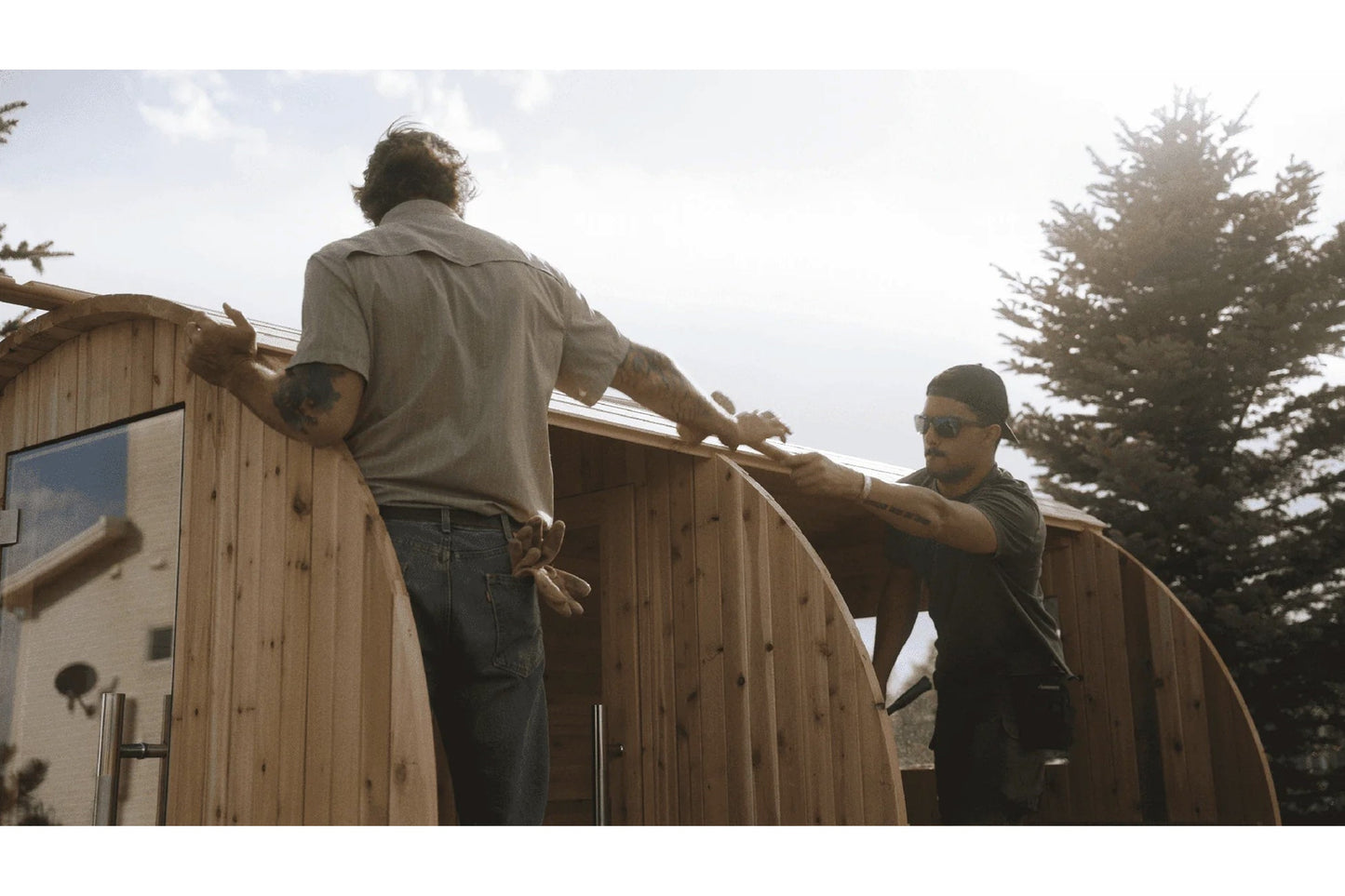 Two men assembling a wooden barrel sauna outdoors, with one wearing a beige shirt and the other in dark clothing and sunglass