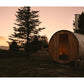 Wooden barrel sauna with metal bands and chimney at sunset, surrounded by evergreen trees and mountains in background