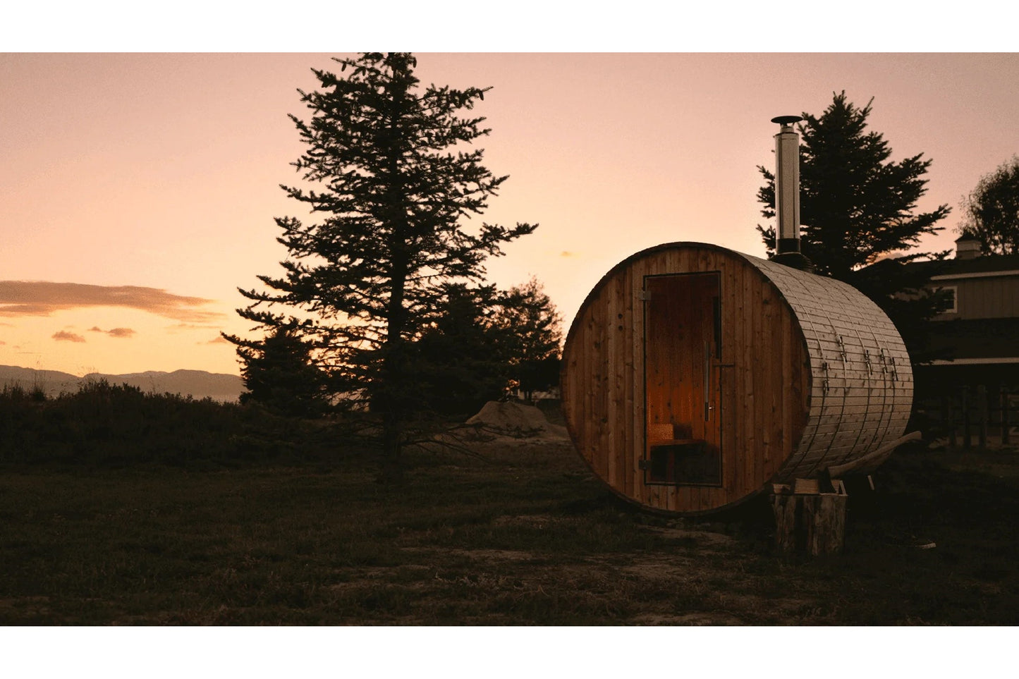 Wooden barrel sauna with metal bands and chimney at sunset, surrounded by evergreen trees and mountains in background