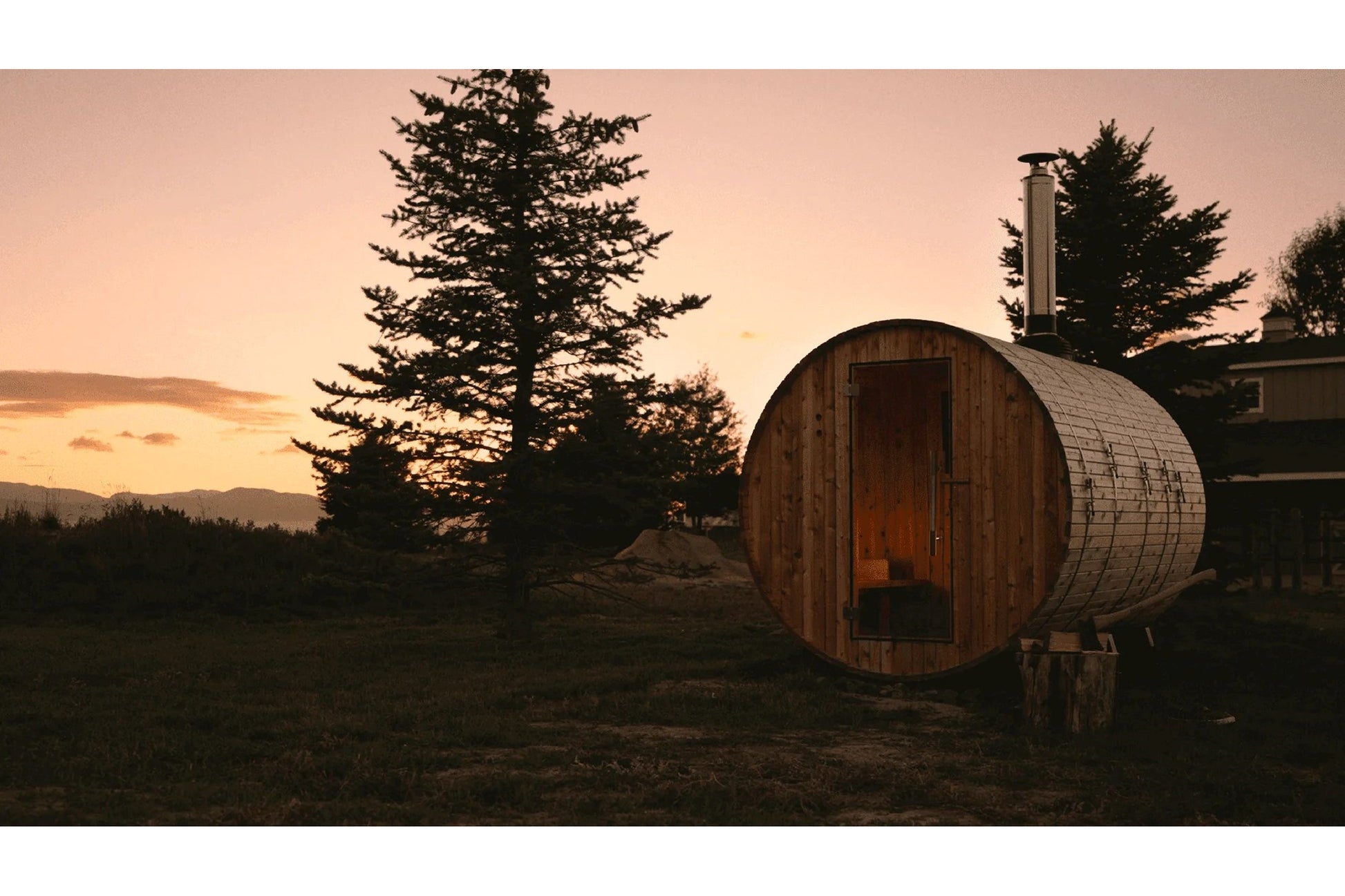 Wooden barrel sauna with metal bands and chimney at sunset, surrounded by evergreen trees and mountains in background
