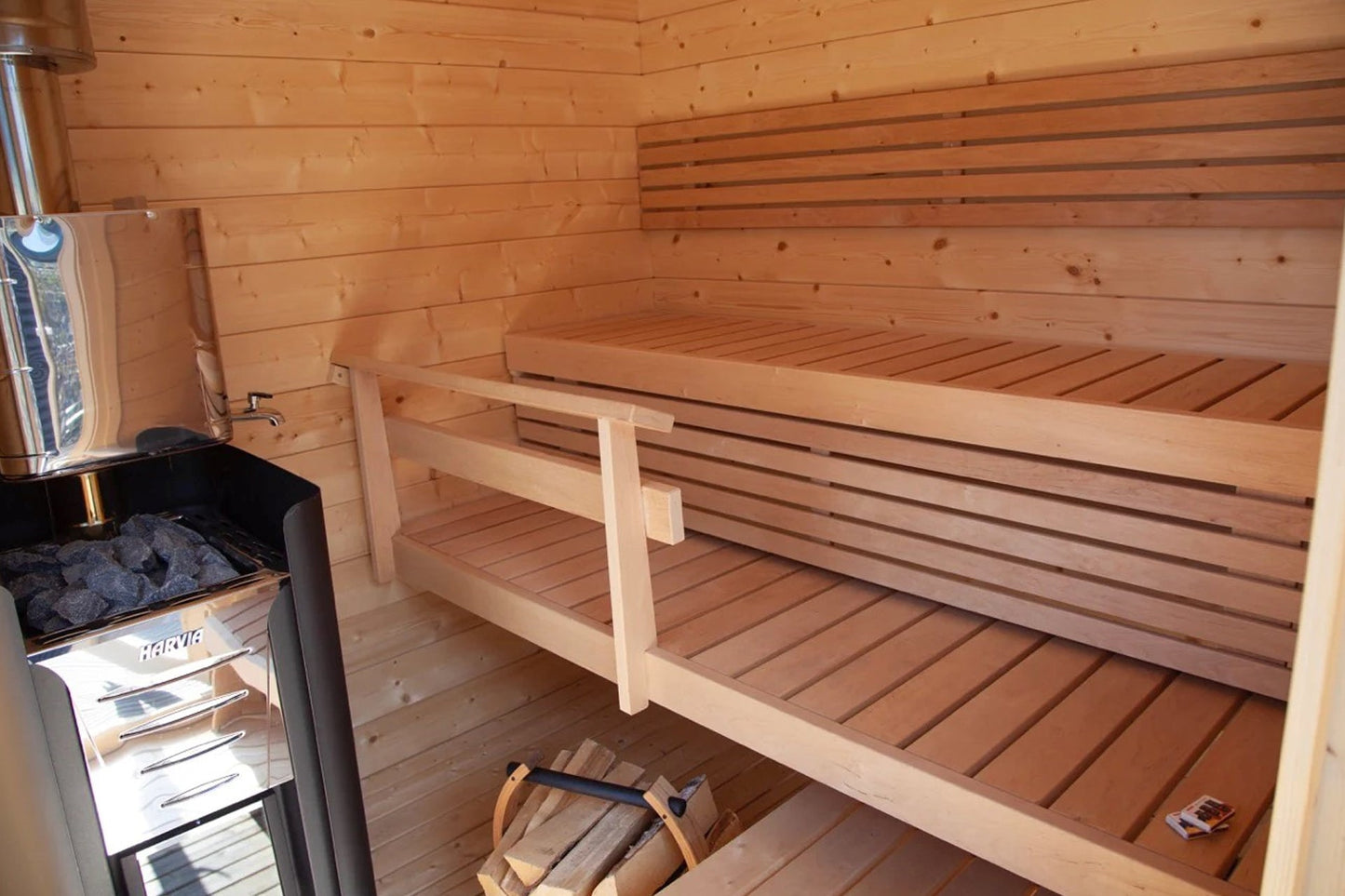 Interior view of wooden sauna cabin with two-tiered benches, Harvia stove with black stones, and light pine construction