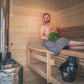 Man using gray towel wrap sitting on wooden sauna bench holding fresh eucalyptus leaves inside wood-paneled sauna cabin