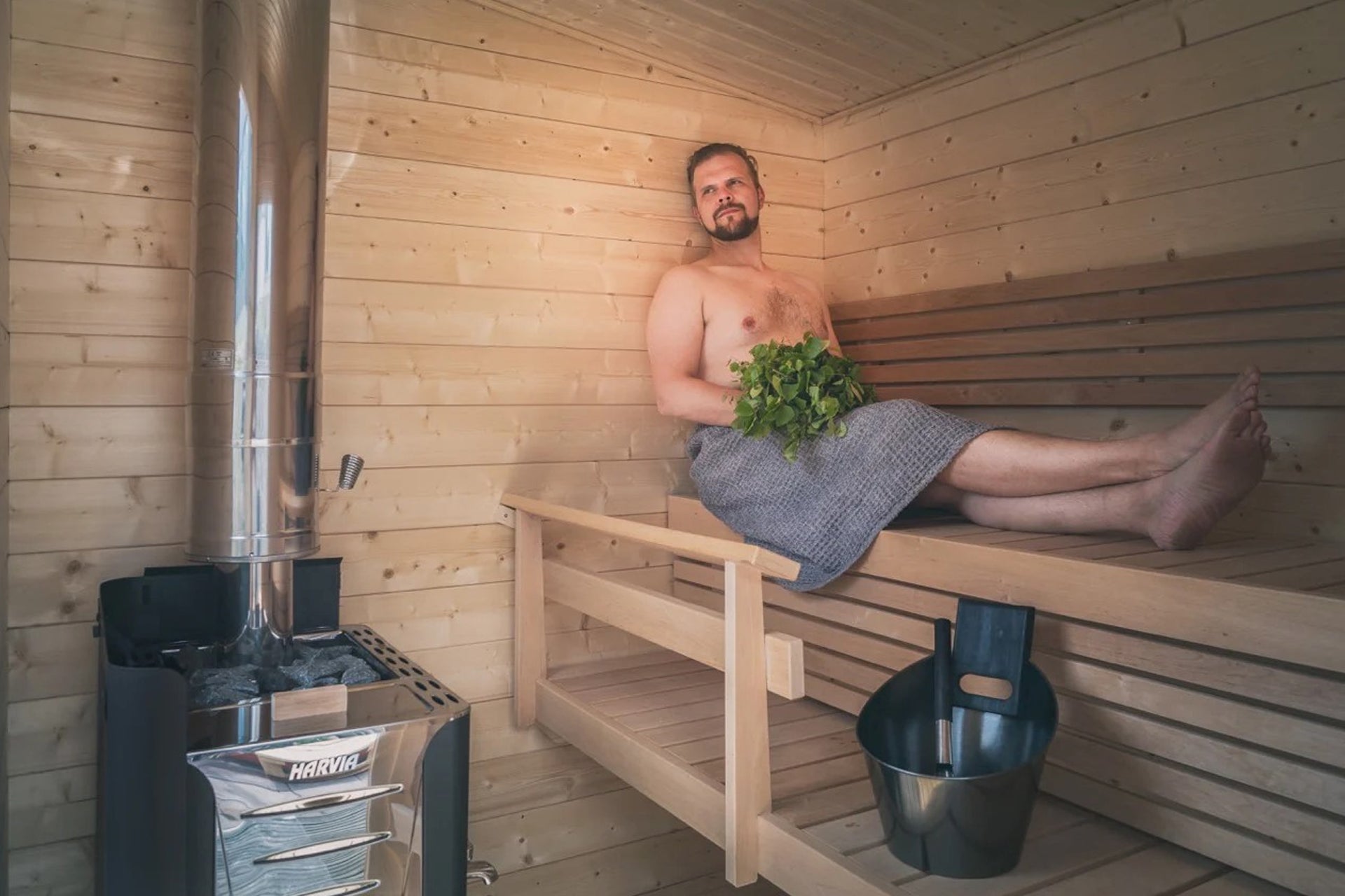 Man using gray towel wrap sitting on wooden sauna bench holding fresh eucalyptus leaves inside wood-paneled sauna cabin