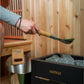 Person holding wooden-handled sauna ladle above Harvia black sauna heater bucket filled with stones in wooden sauna room