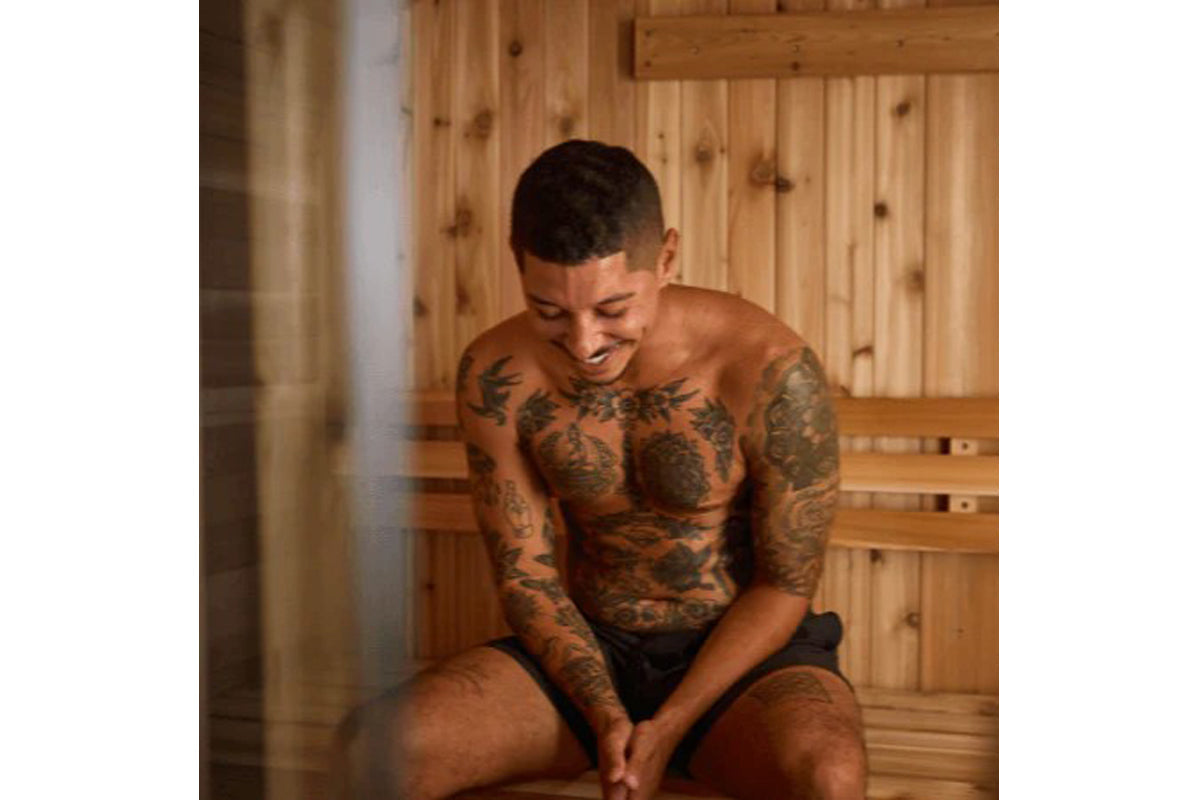 Shirtless man with extensive chest and arm tattoos sitting in wooden sauna interior, worn by model
