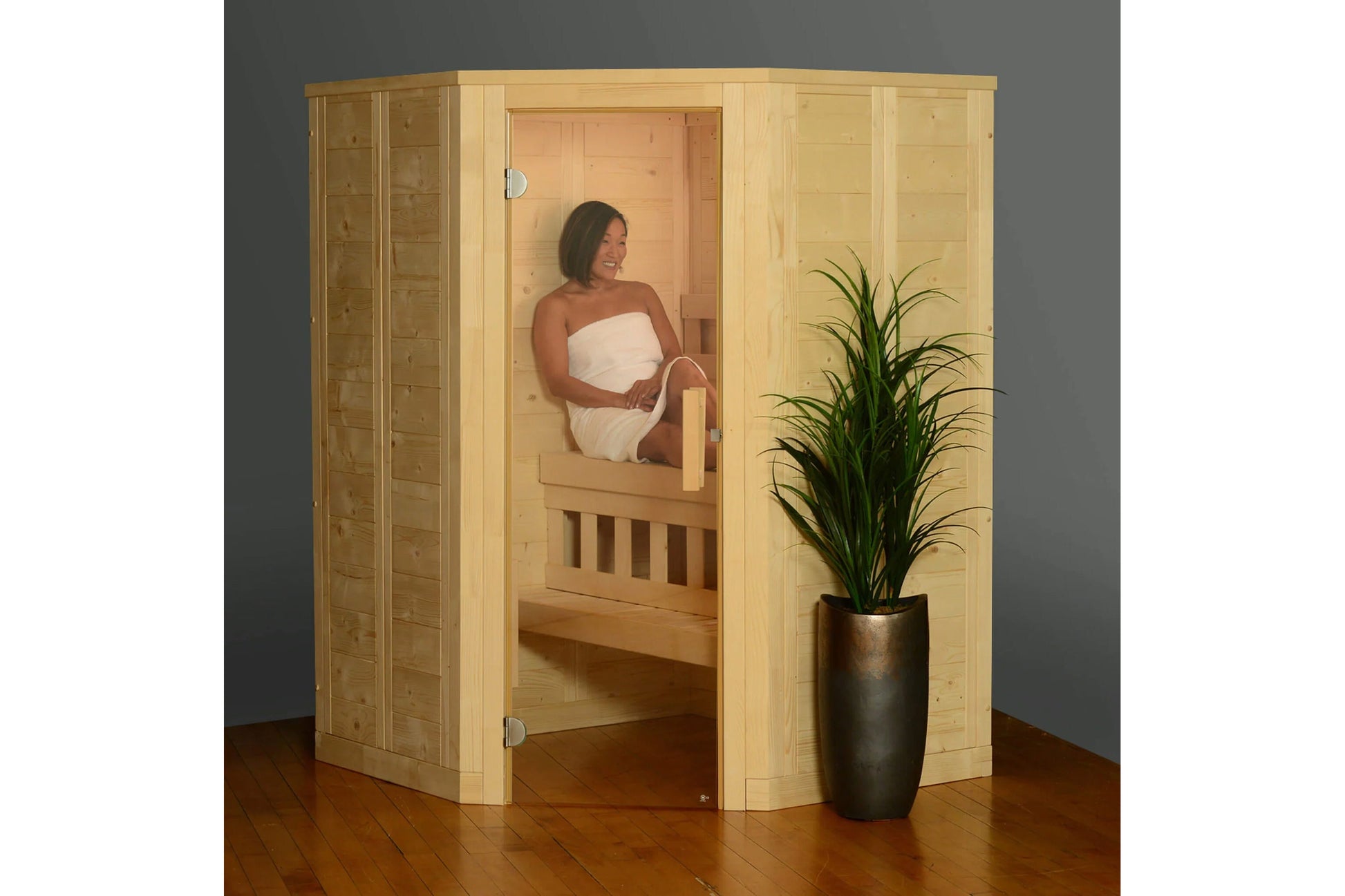 Light wood corner sauna with glass door, woman seated inside wrapped in white towel, potted plant beside sauna on wooden floo